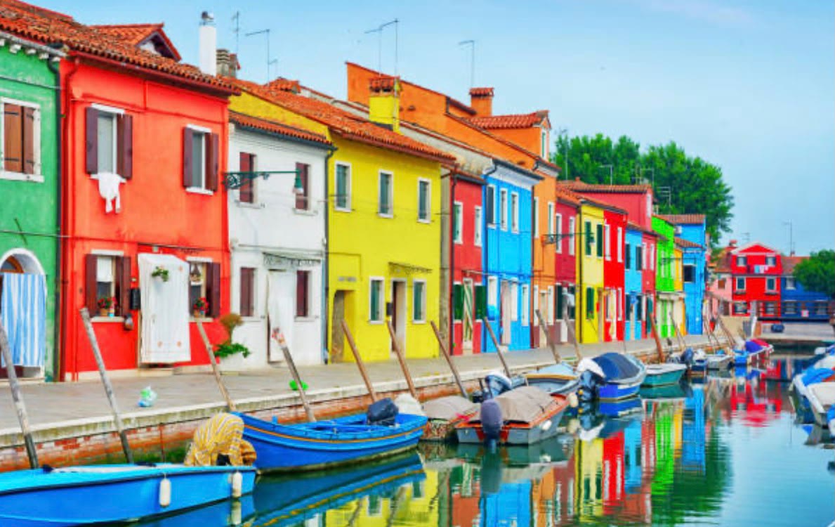 The brightly coloured fishermen's houses of Burano mirrored in a still canal, moored boats in the foreground